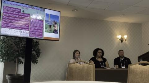 Three people on a panel with TV monitor displaying research stats in upper left corner of photo