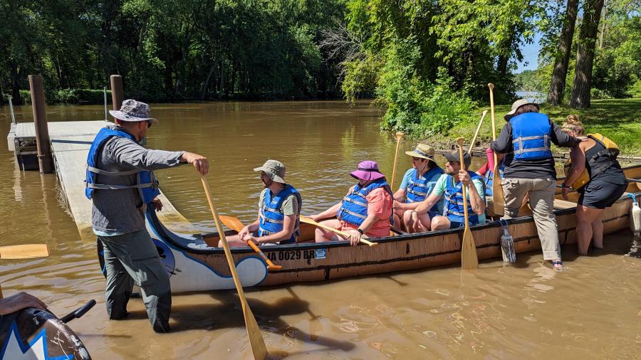 Adults with life jackets and paddles in a long canoe preparing to launch into a river